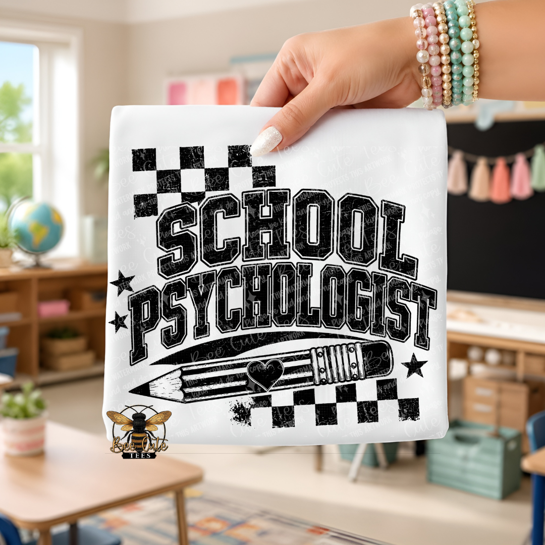 A hand holds up a white sweatshirt featuring a black graphic that reads 'SCHOOL PSYCHOLOGIST' with a pencil, stars, and checkerboard accents, set against a blurred classroom background.