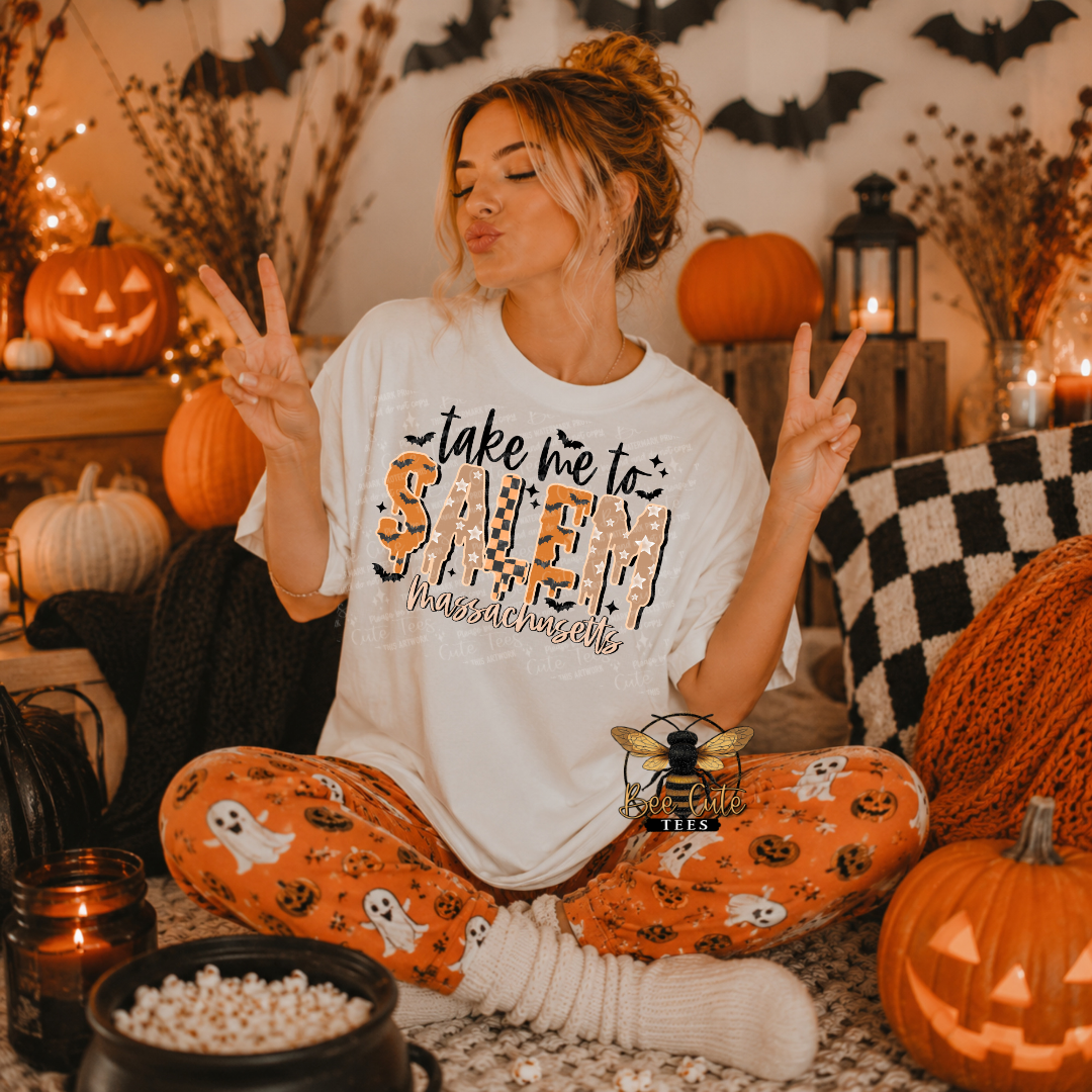 A woman sits cross-legged in a cozy Halloween-themed room, wearing a white graphic t-shirt that says 'take me to SALEM Massachusetts' and orange pajama pants with a ghost and pumpkin print. She holds up peace signs, surrounded by pumpkins, candles, and bats.