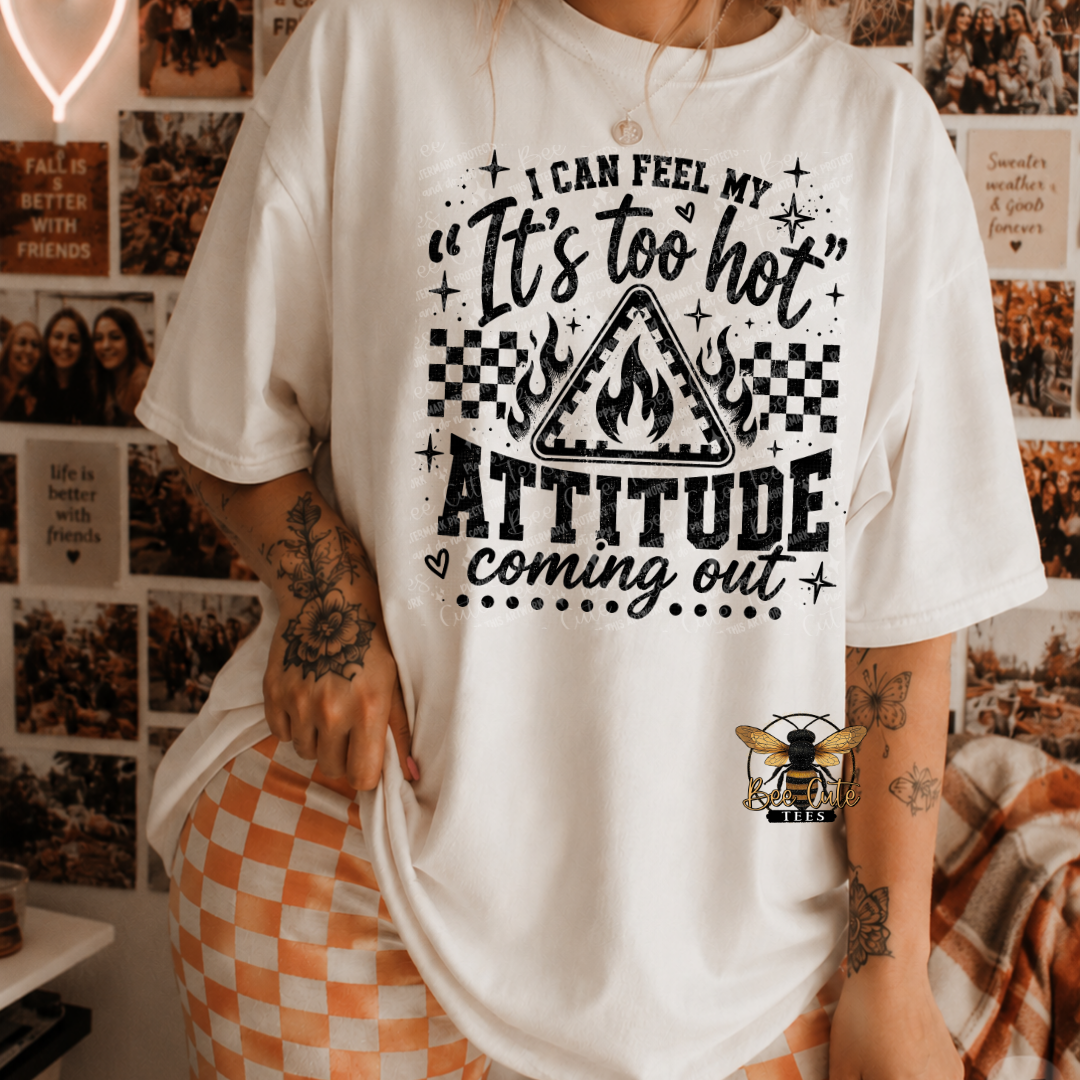 A person wearing a white graphic T-shirt with black text reading “I CAN FEEL MY ‘It’s too hot’ ATTITUDE coming out” and a flame triangle graphic, paired with orange-and-white checkered shorts in a decorated indoor room.