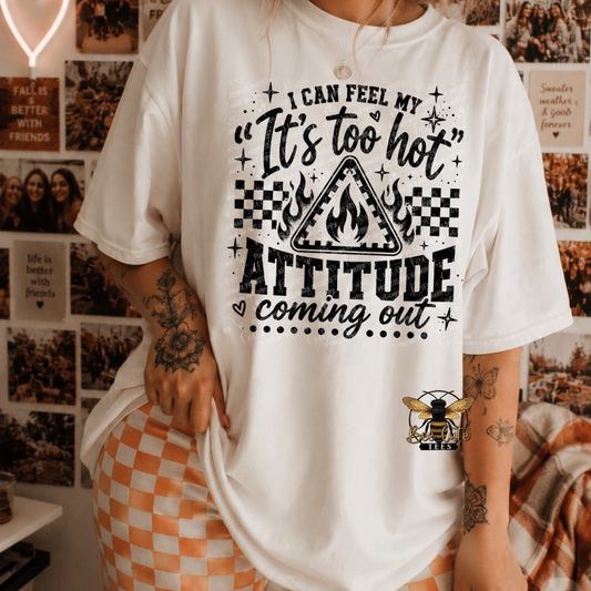 A person wearing a white graphic T-shirt with black text reading “I CAN FEEL MY ‘It’s too hot’ ATTITUDE coming out” and a flame triangle graphic, paired with orange-and-white checkered shorts in a decorated indoor room.