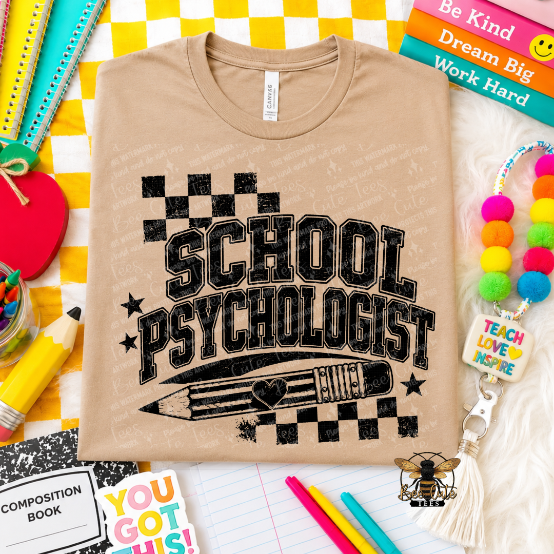 A beige graphic t-shirt featuring the words 'SCHOOL PSYCHOLOGIST' in distressed black lettering with a pencil and checkered border graphic. The shirt is displayed in a styled flat-lay arrangement with school supplies, books, and a colorful pom-pom keychain.