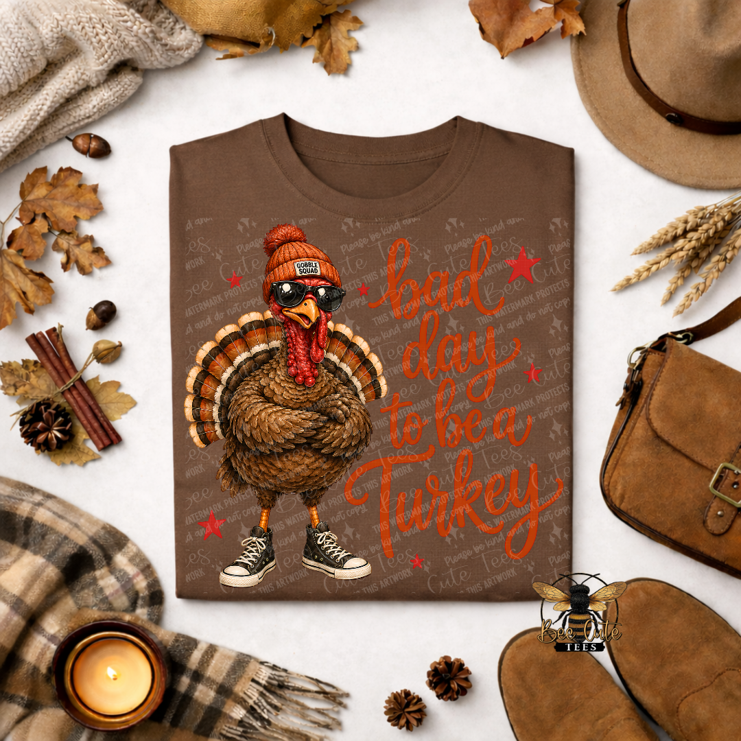 A brown graphic t-shirt featuring a cartoon turkey wearing sunglasses and a beanie with the text 'bad day to be a Turkey' in orange script. The shirt is displayed in a flat-lay arrangement with autumn-themed props including a hat, shoes, a candle, and leaves on a white background.