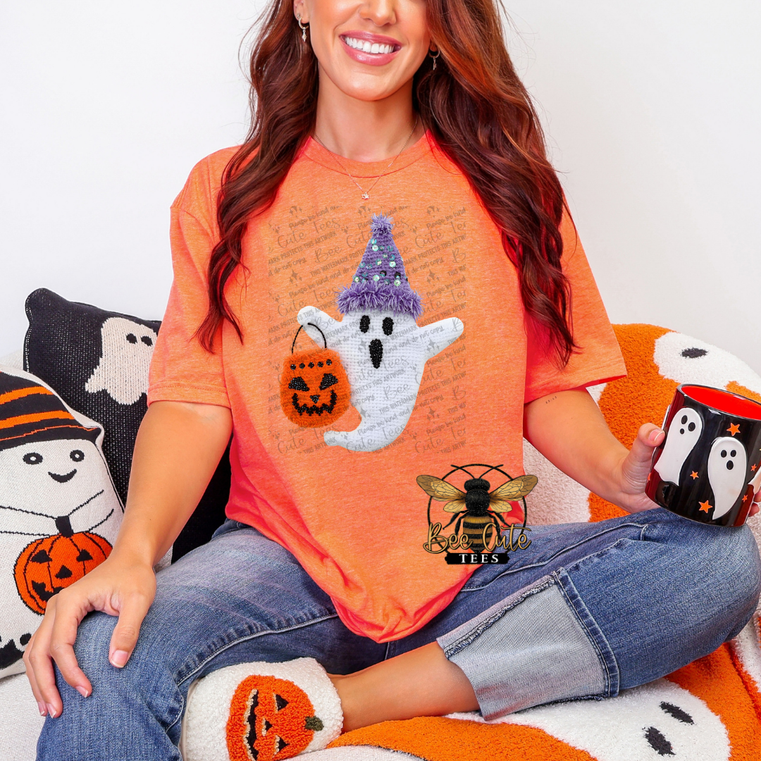 A woman wearing a bright orange t-shirt with a Halloween-themed graphic of a ghost in a purple hat holding a pumpkin bag, sitting on a cozy white and orange couch and holding a matching black and white mug with a ghost design.