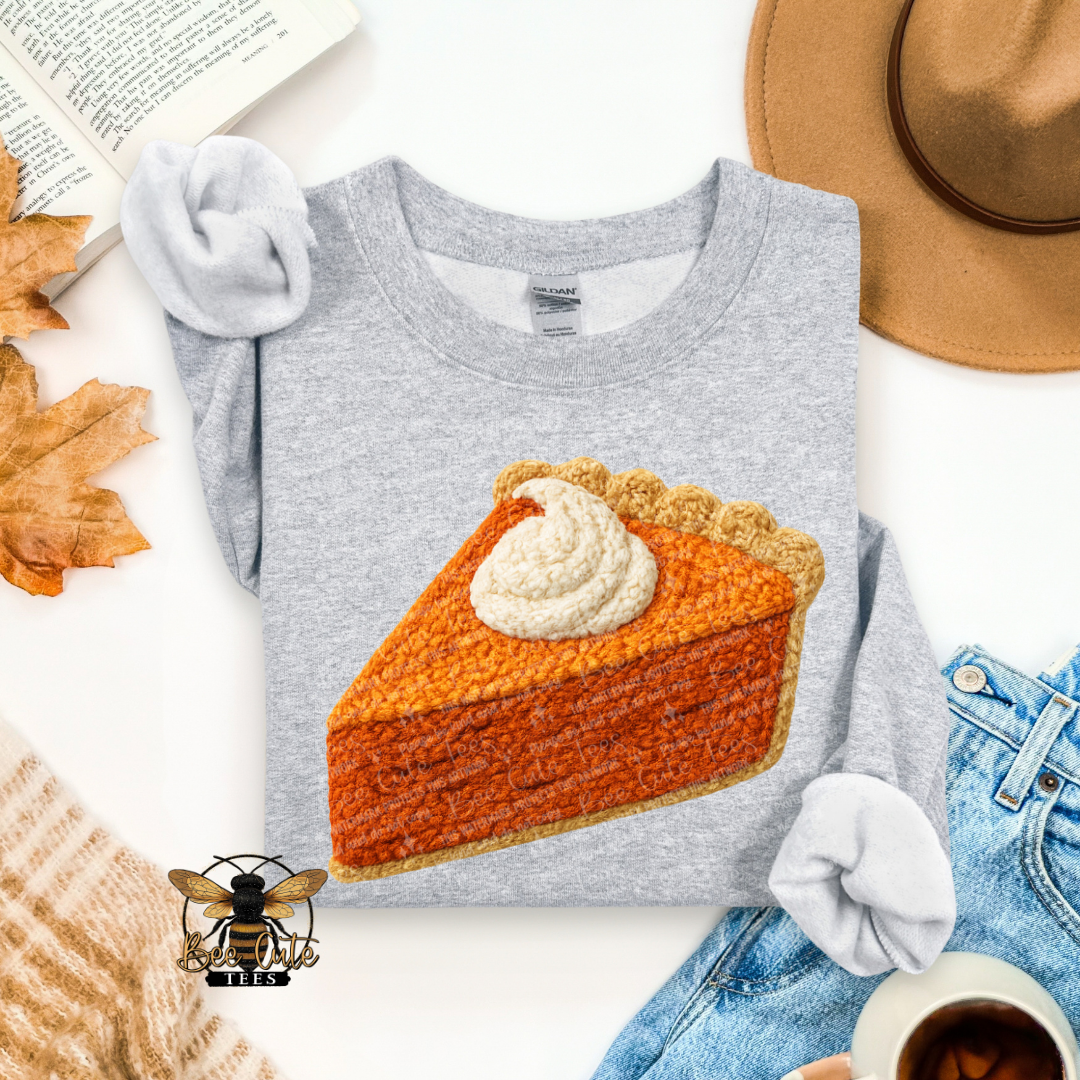 A heather gray long-sleeve t-shirt with a large crochet-knit style graphic of a pumpkin pie slice on the front, styled in a flat lay with a tan hat, blue jeans, a book, autumn leaves, a knit sweater, and a cup of coffee.