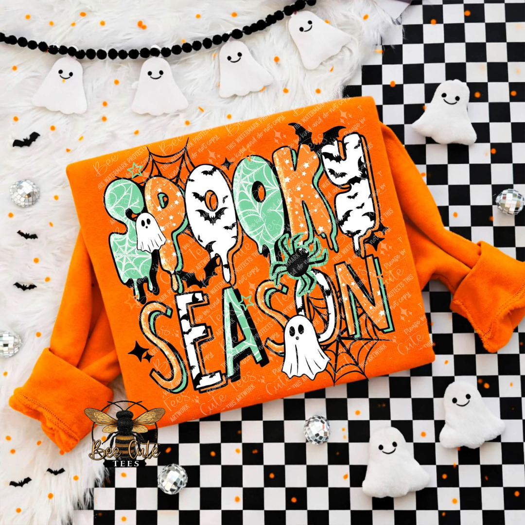 An orange long-sleeve sweatshirt laid flat on a Halloween-themed background. The front has a large decorative graphic reading “Spooky Season” in mixed white, mint green, black, and orange lettering with ghosts, bats, spiders, and spiderwebs.