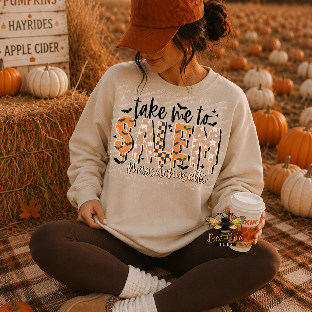 A woman sits cross-legged on a plaid blanket in a fall-themed pumpkin field, wearing a cream sweatshirt with a graphic that reads 'take me to SALEM Massachusetts' in orange, black, and white. She is also wearing a rust-orange cap and brown leggings, holding a coffee cup.