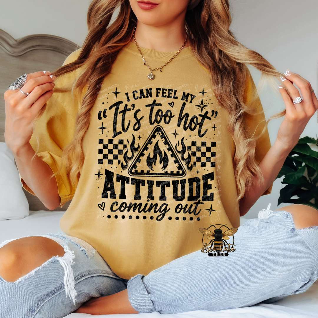 A woman wearing a mustard yellow graphic t-shirt with a black text design that reads 'I CAN FEEL MY "It's too hot" ATTITUDE coming out' with a flame warning sign graphic. She is sitting on a bed wearing distressed light blue jeans.