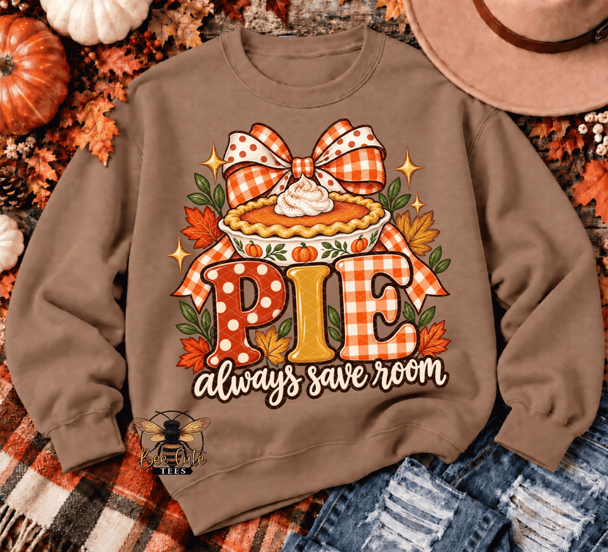 A brown crewneck sweatshirt featuring a fall-themed pie graphic with the text 'PIE always save room'. The sweatshirt is styled in a flat-lay arrangement with autumn props including pumpkins, a cowboy hat, a plaid blanket, and denim jeans.