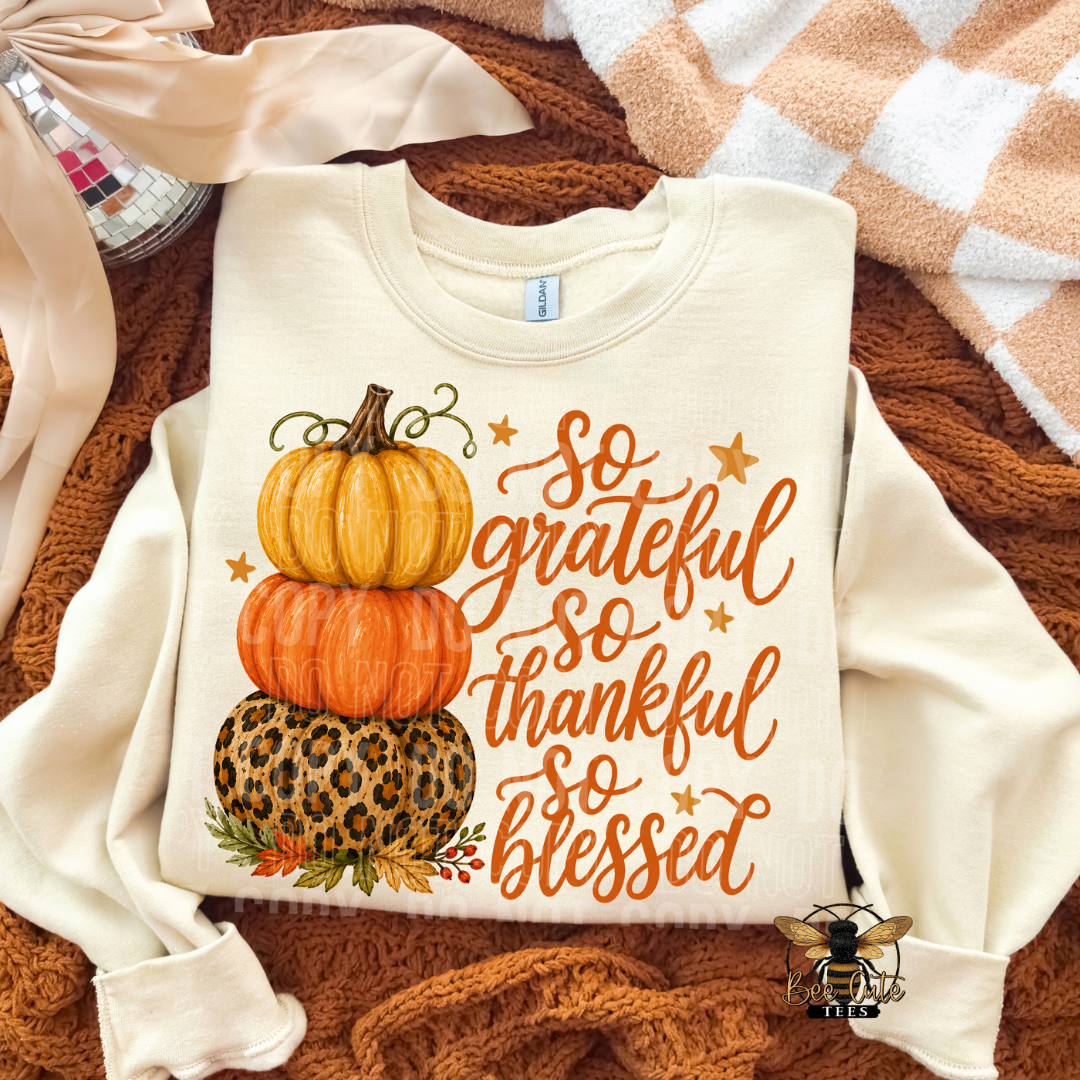 An off-white long-sleeve sweatshirt laid flat on a brown knit blanket, featuring a stacked pumpkin graphic and the phrase 'so grateful so thankful so blessed' in orange script. A small bee logo appears in the lower right corner.