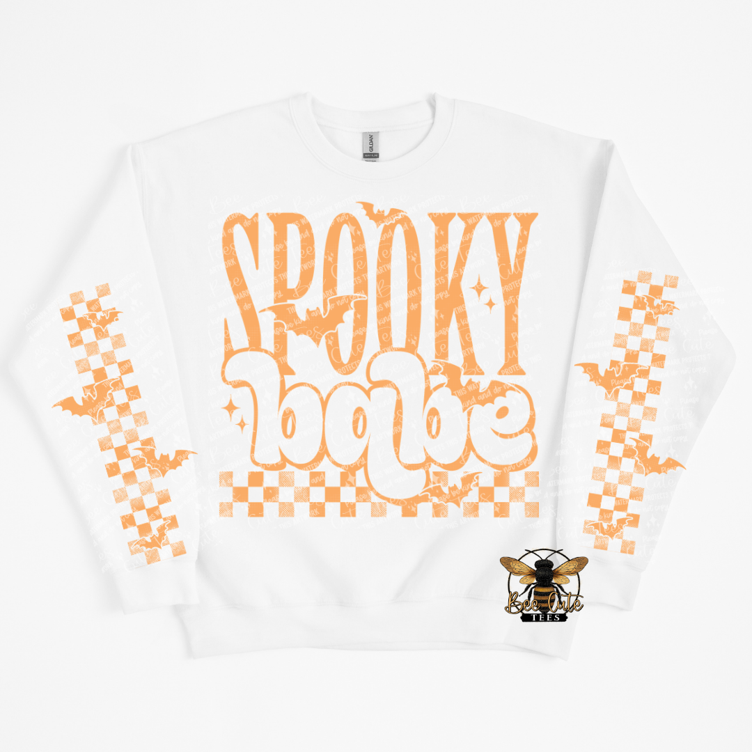 A white crewneck sweatshirt featuring a distressed orange graphic that reads 'Spooky babe' with bats and stars, orange checkered patterns on both sleeves, and a small bee logo on the lower right front.