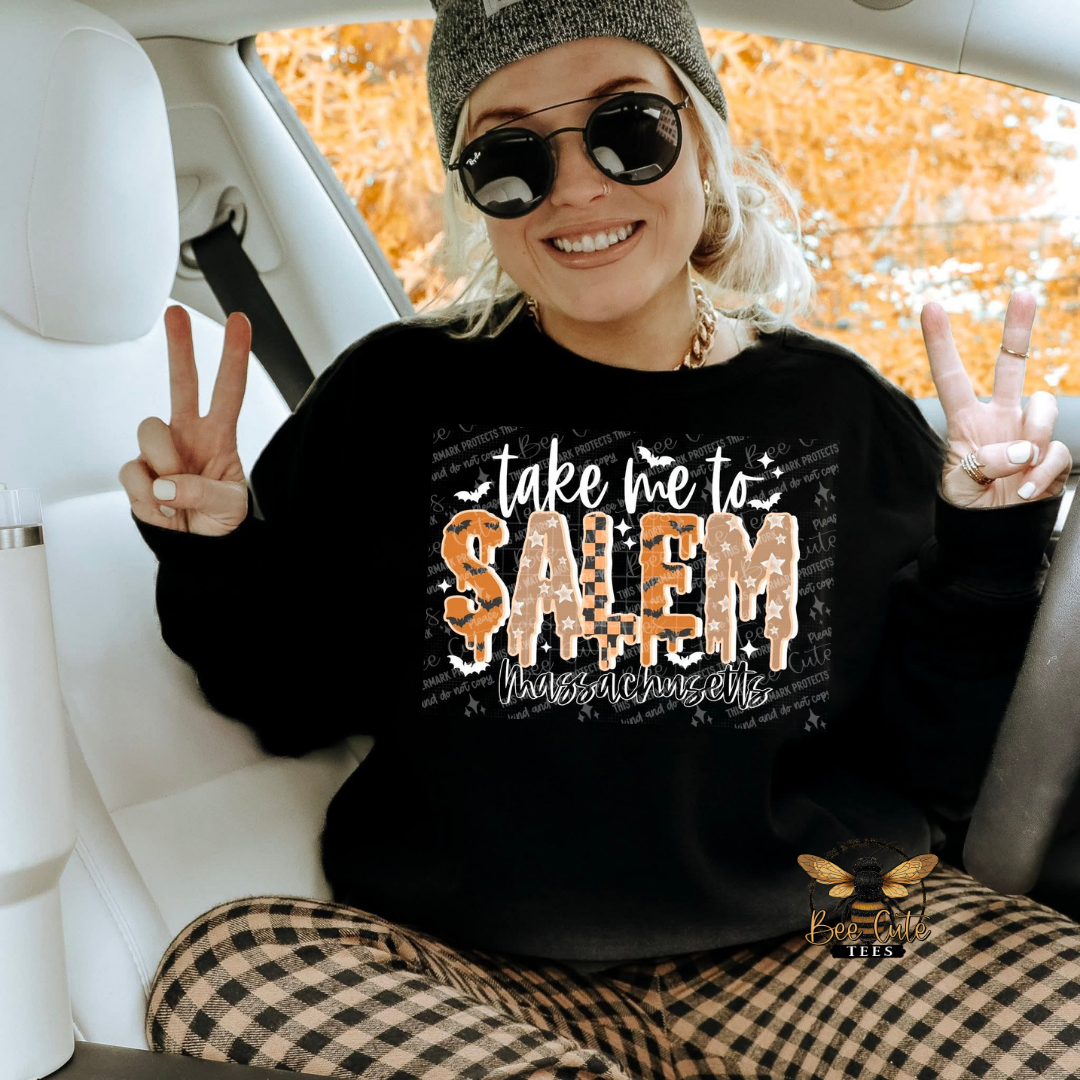 A woman sitting inside a car wearing a black crewneck sweatshirt with a 'take me to SALEM Massachusetts' graphic, paired with brown and black plaid pants. She is smiling, wearing sunglasses, and making peace signs.