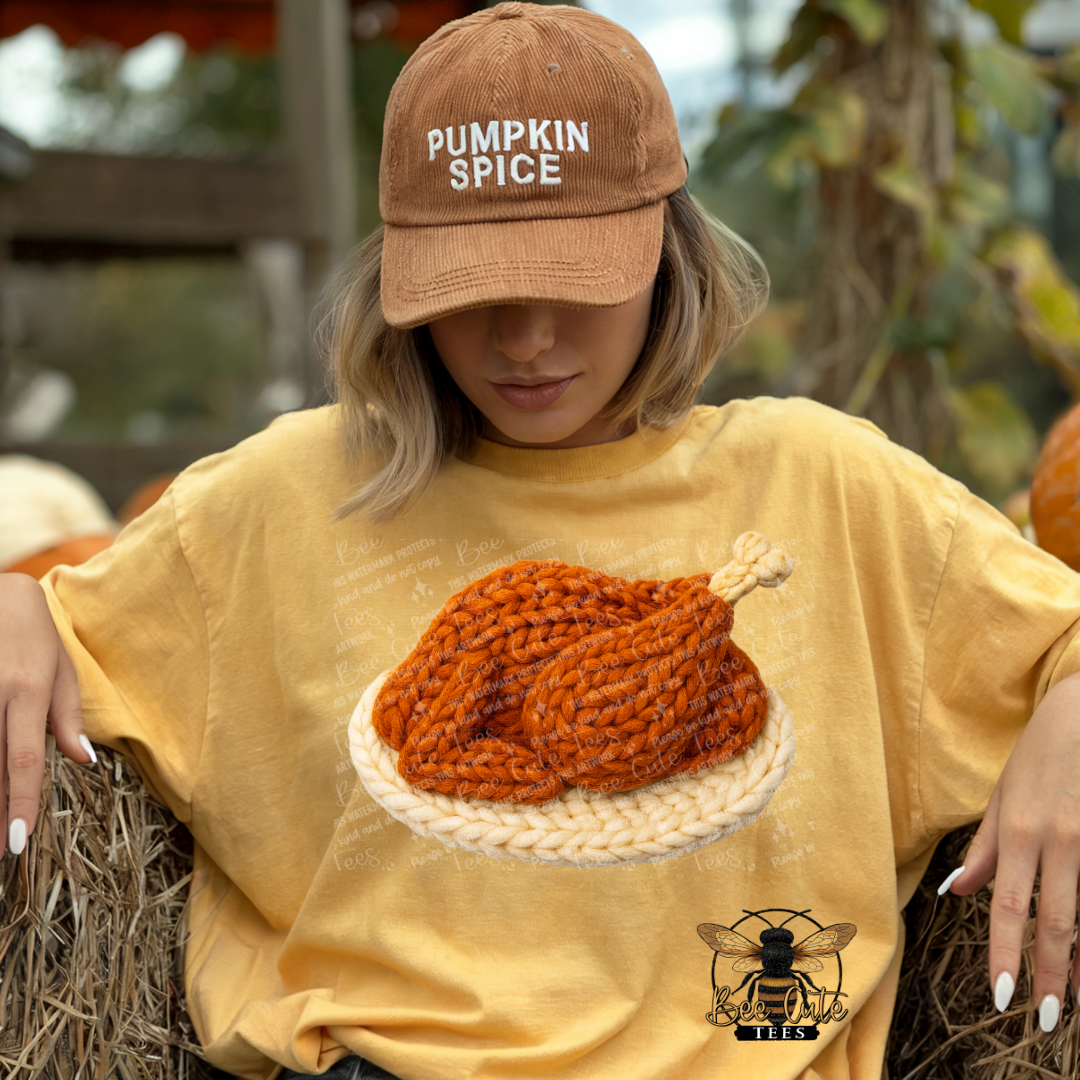 A woman wearing a tan 'PUMPKIN SPICE' baseball cap and a yellow graphic t-shirt with a large knit turkey design, sitting on hay bales in a pumpkin patch.