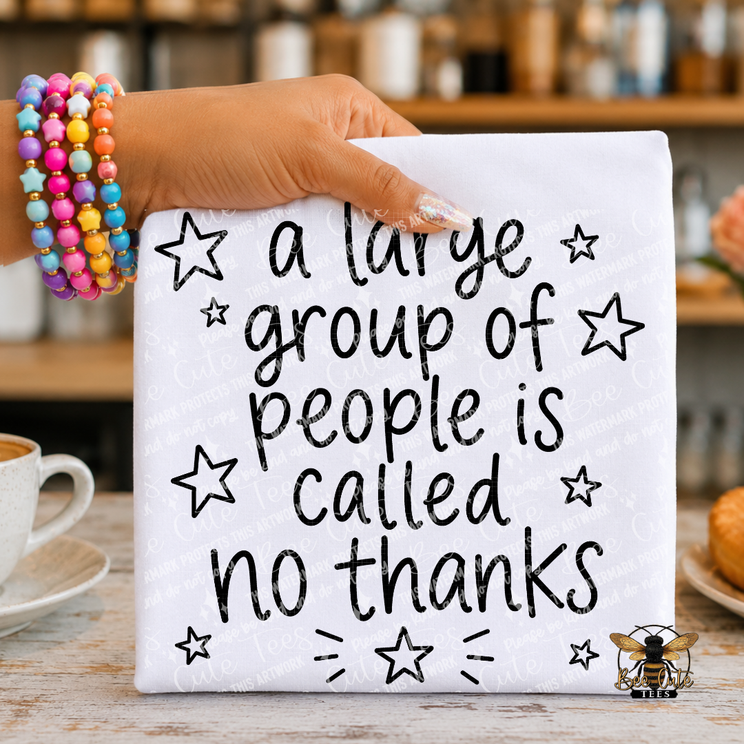 A hand wearing colorful beaded bracelets holds a white folded t-shirt with black handwritten-style text reading “a large group of people is called no thanks,” decorated with black outlined stars. The shirt is photographed in a real indoor setting with a blurred kitchen or cafe background, a cup at left, and a pastry at right.