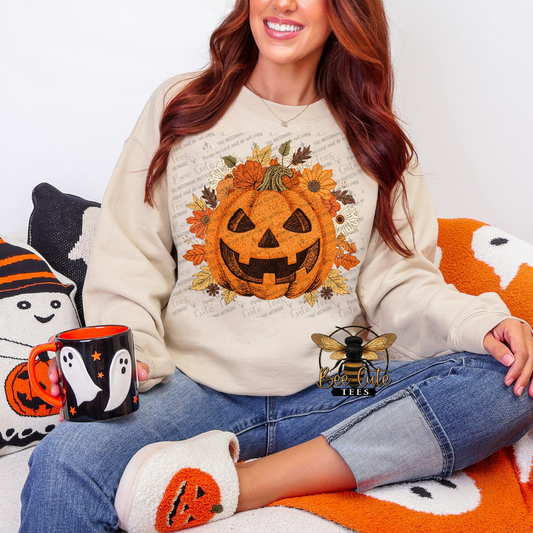 A woman wearing a cream-colored crewneck sweatshirt with a large orange pumpkin graphic surrounded by autumn leaves and flowers. She is sitting on a cozy blanket with Halloween-themed pillows, holding a black mug with a ghost design.