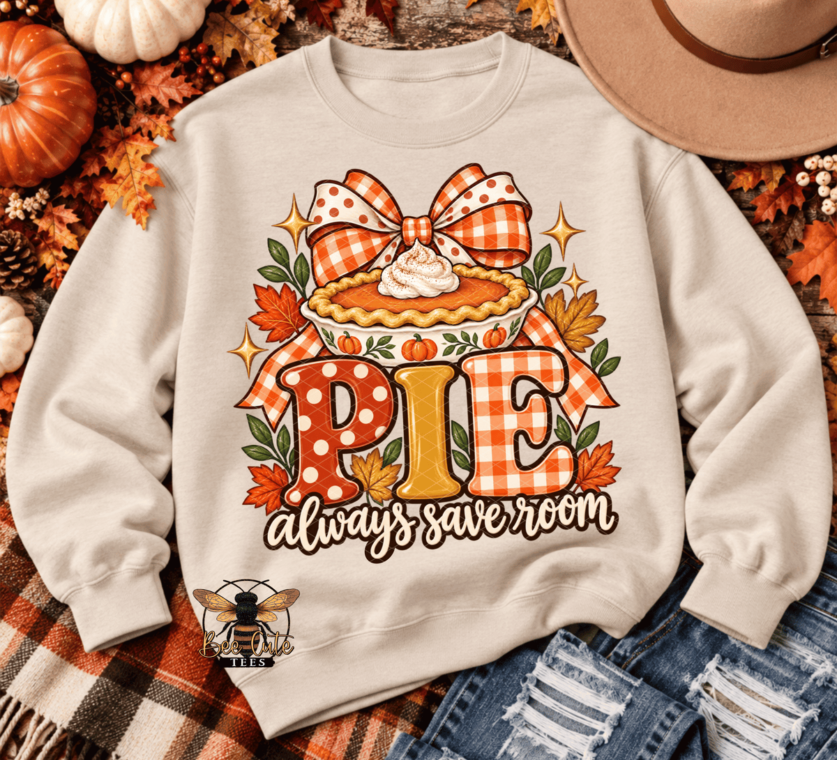 A cream-colored crewneck sweatshirt featuring a festive autumn 'Pie Always Save Room' graphic, styled in a flat-lay with fall leaves, pumpkins, a cowboy hat, and denim jeans.