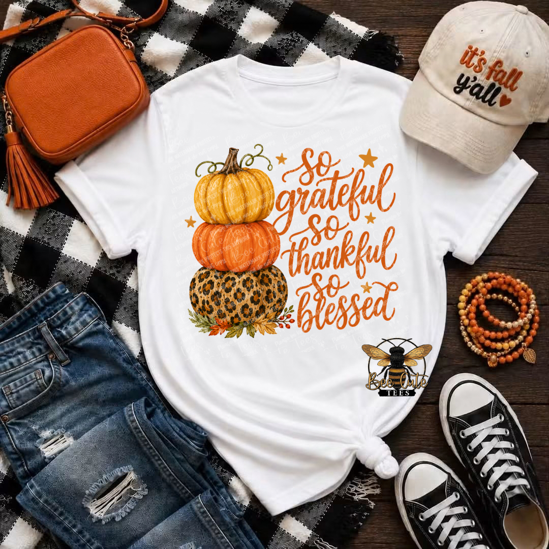 A white graphic t-shirt with a stacked autumn pumpkin design and the text 'so grateful so thankful so blessed' in orange script, styled in a flat lay with a black-and-white plaid blanket, orange crossbody bag, distressed blue jeans, beige 'it's fall y'all' cap, beaded bracelet, and black-and-white sneakers.