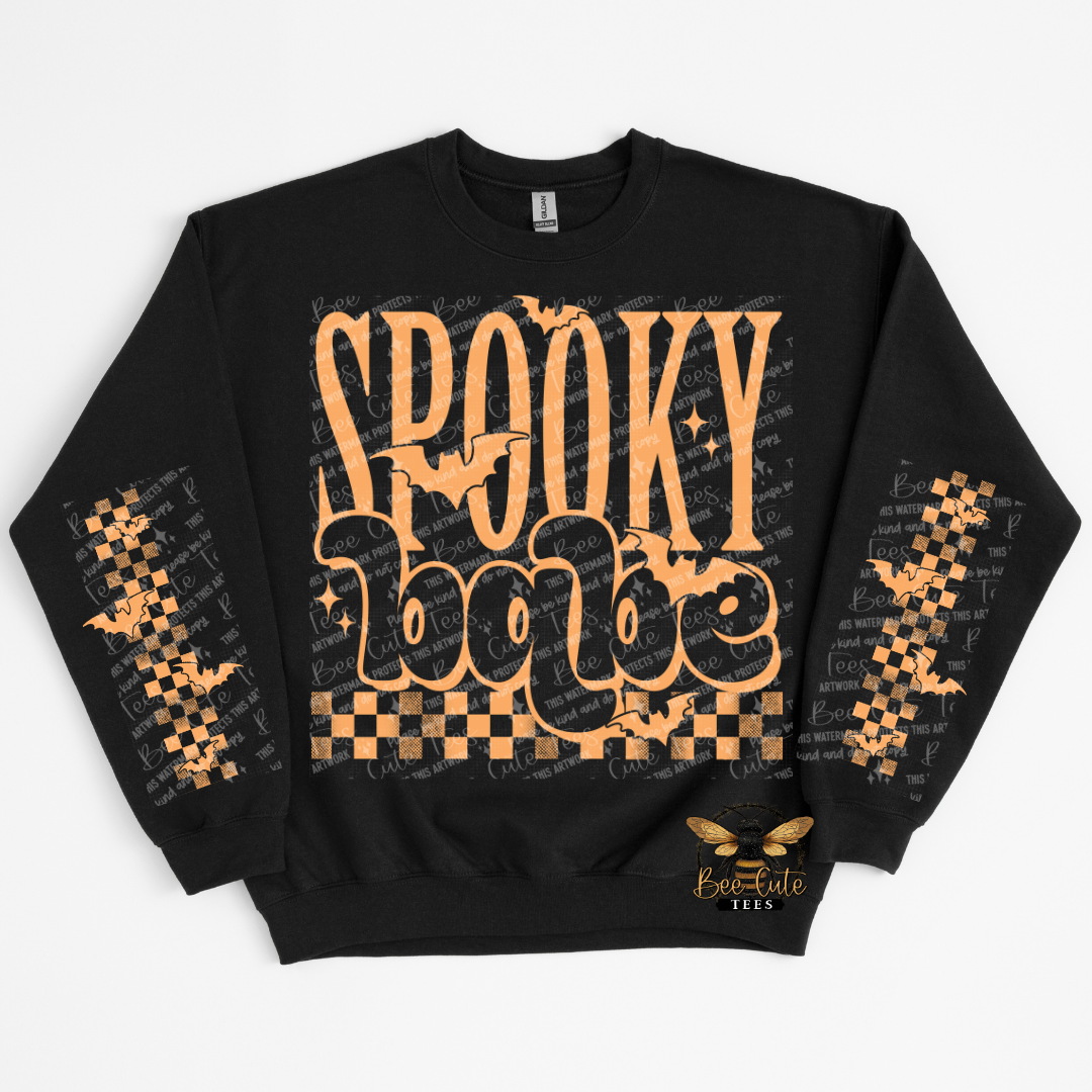 A black crewneck sweatshirt featuring a 'Spooky Babe' graphic in orange on the front, decorated with orange bats and checkered patterns on the sleeves, with a bee logo on the lower right front.