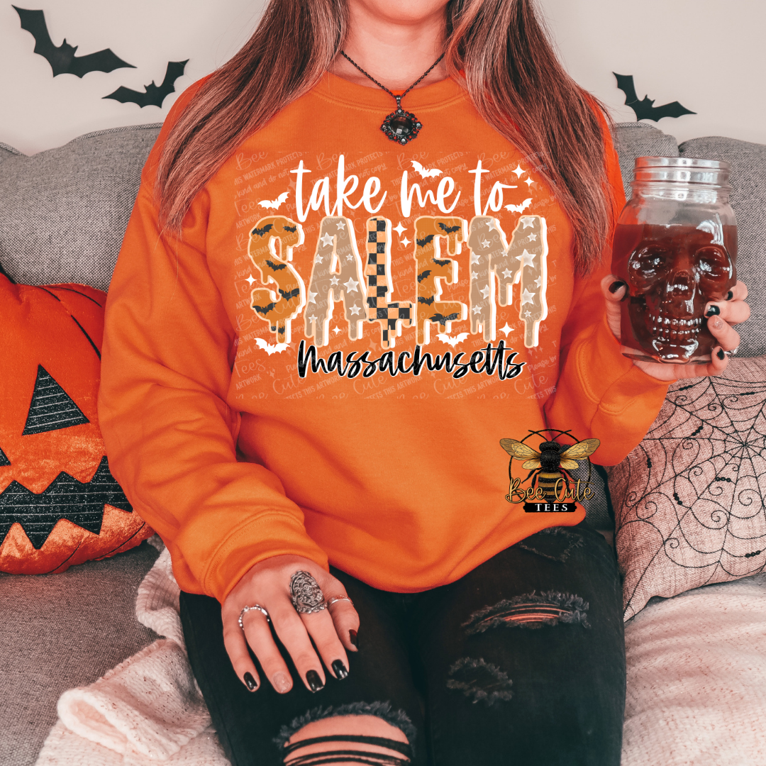 A woman wearing an orange sweatshirt with a Halloween-themed graphic that reads 'take me to SALEM Massachusetts' in a spooky font. She is sitting on a couch holding a skull-shaped mason jar, with Halloween decorations like a pumpkin pillow and spiderweb pillow in the background.