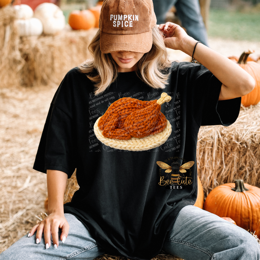 A woman sitting outdoors on hay bales wears a black graphic T-shirt featuring a large knit-style turkey graphic and the words 'Be Cute Tees,' along with a brown 'Pumpkin Spice' cap.