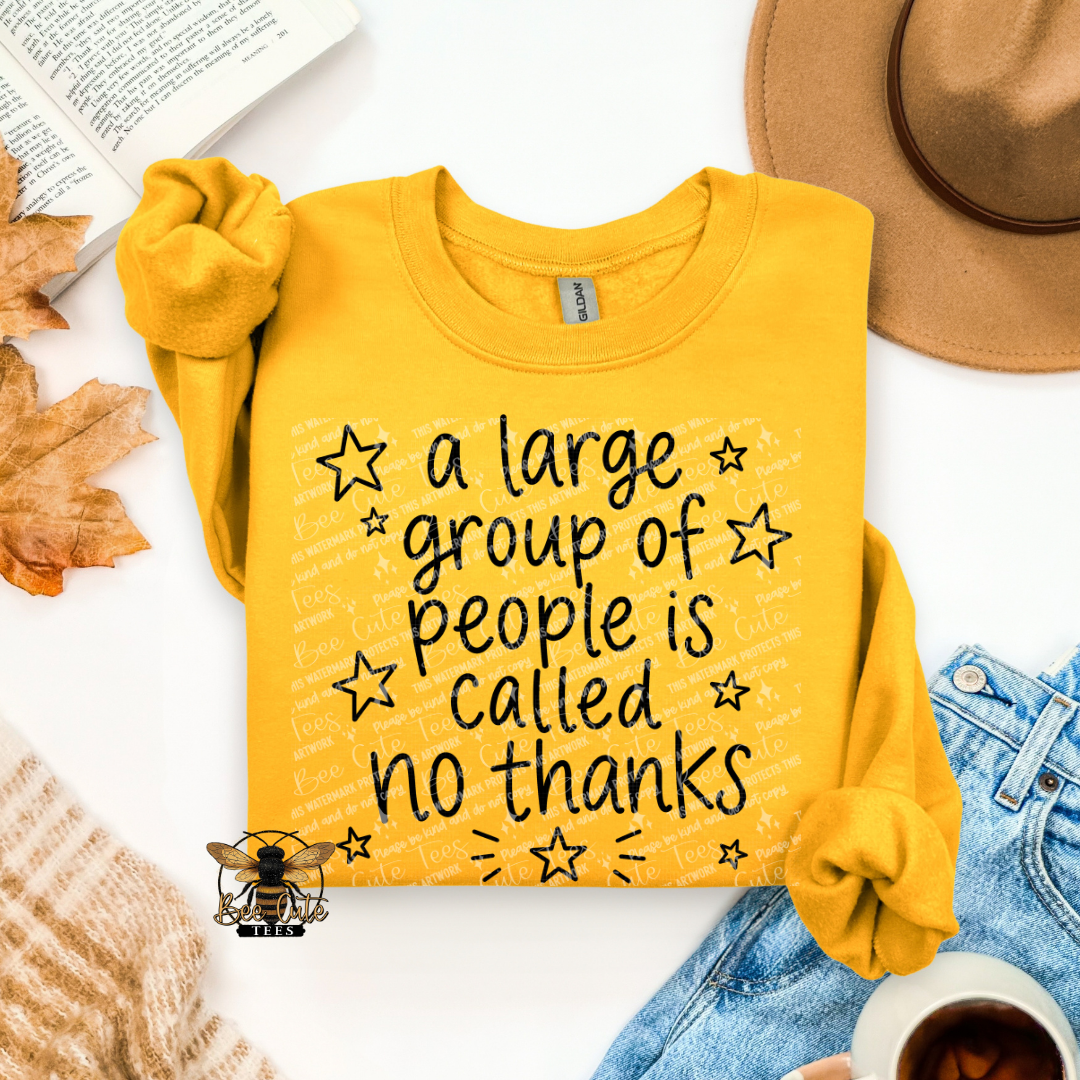 A folded yellow long-sleeve T-shirt with rolled sleeves, printed with the phrase 'a large group of people is called no thanks' in black script with star accents. The shirt is styled in a flat lay with a tan hat, blue jeans, a cup of coffee, a book, and autumn leaves on a white background.
