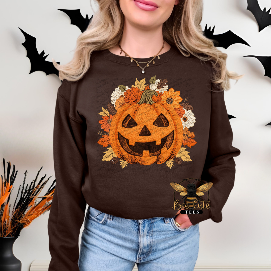 A woman wearing a dark brown crewneck sweatshirt with a large Halloween graphic of a jack-o'-lantern surrounded by autumn leaves and flowers. The sweatshirt features a 'bee cute tees' watermark logo on the bottom right.