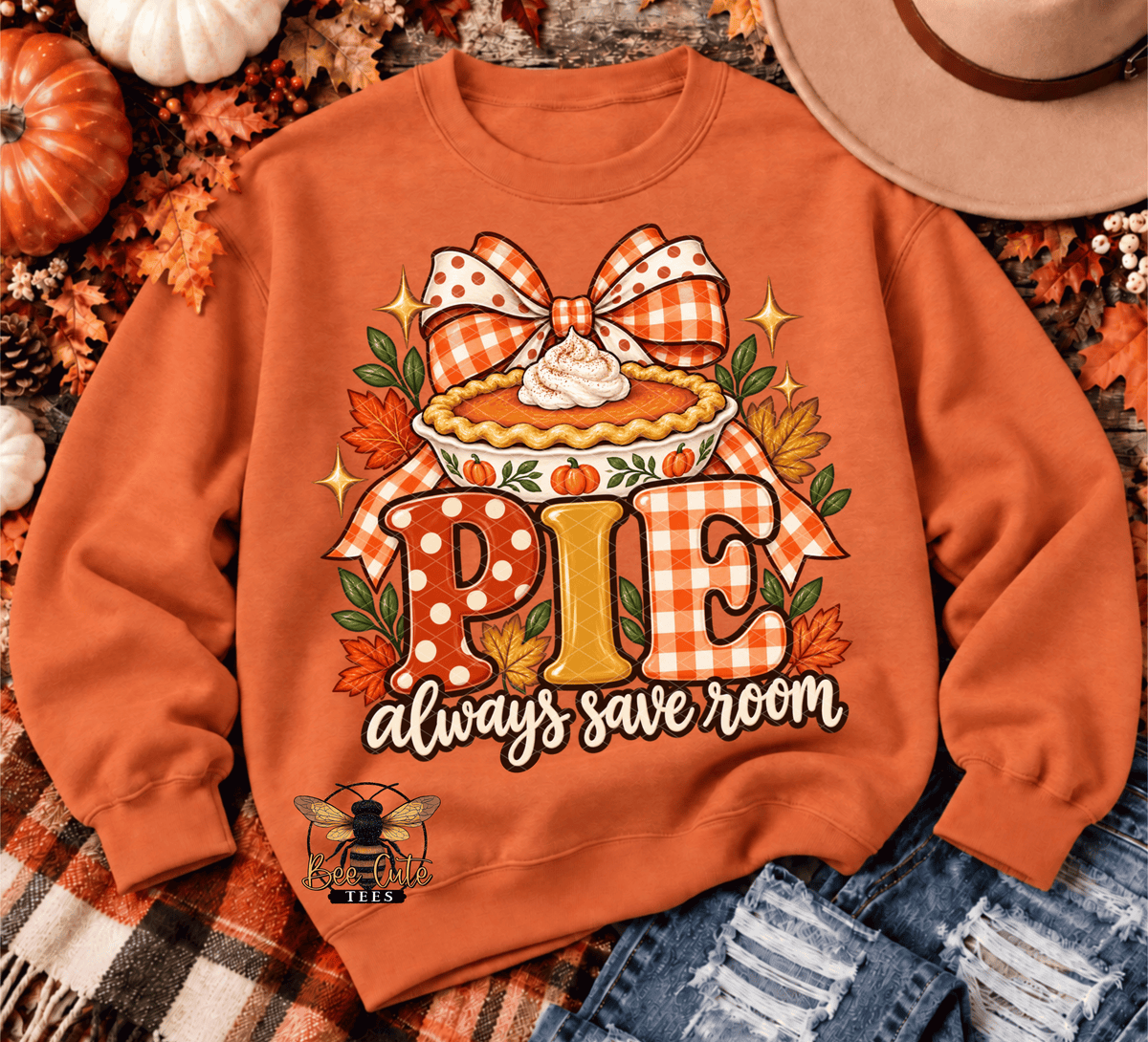 An orange crewneck sweatshirt featuring a fall-themed pie graphic with the text 'PIE always save room'. The sweatshirt is styled in a flat lay with autumn props including a small pumpkin, a cowboy hat, denim, and a plaid blanket.