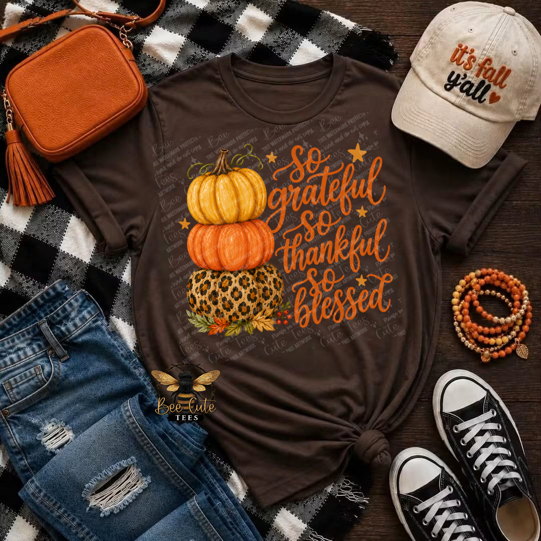 A brown graphic t-shirt featuring stacked pumpkins and the text 'So grateful SO thankful SO blessed' in orange, styled in a flat lay with a black-and-white plaid blanket, jeans, a brown crossbody bag, a beige 'It's fall y'all' cap, and black-and-white sneakers.