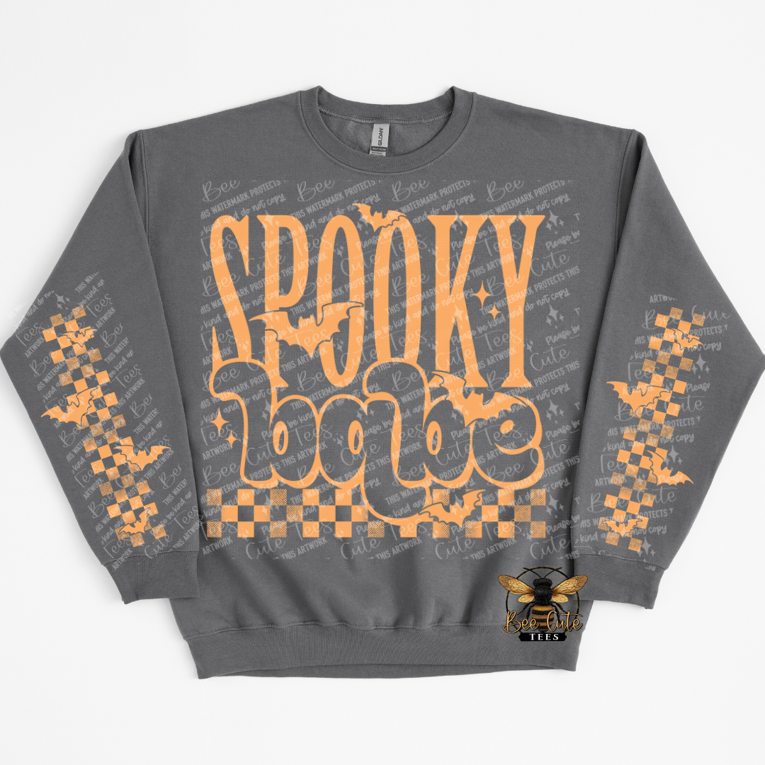 A dark gray crewneck sweatshirt featuring a 'Spooky Babe' graphic in orange on the front, decorated with orange bats and checkered patterns on the sleeves, with a bee logo on the lower right front.