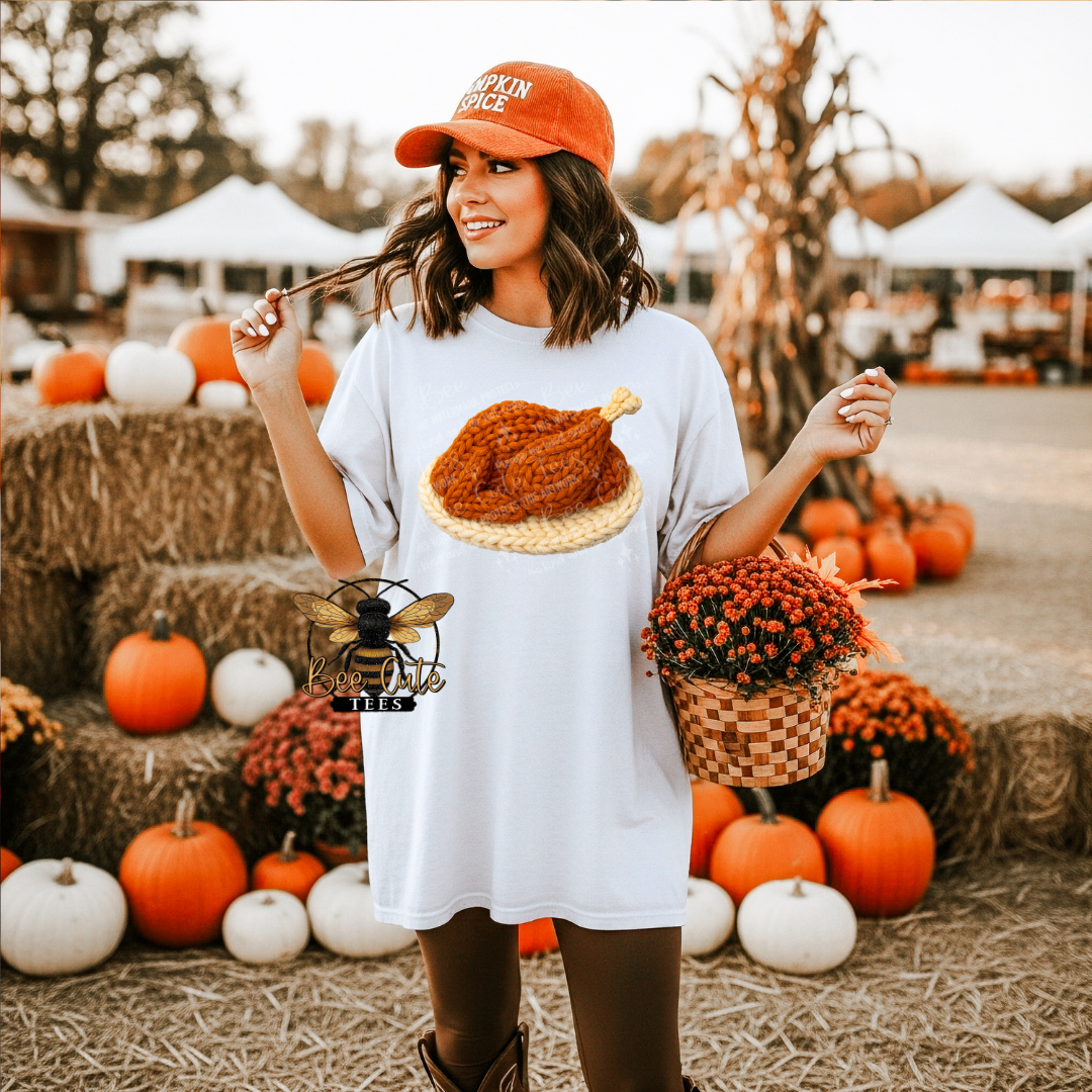 A woman stands outdoors at a fall farm market wearing an oversized white T-shirt dress with a large roasted turkey graphic on the front, an orange baseball cap, and holding pumpkins and flowers. The background shows hay bales, pumpkins, corn, and white market tents.