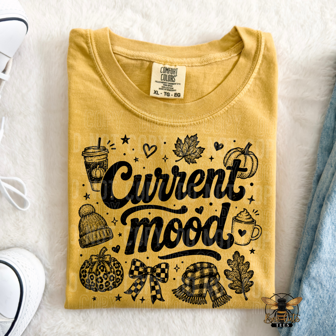 A folded mustard yellow t-shirt with a black graphic reading 'Current mood' surrounded by autumn illustrations, laid flat on a white faux fur surface with partial jeans and a shoe visible at the edges.