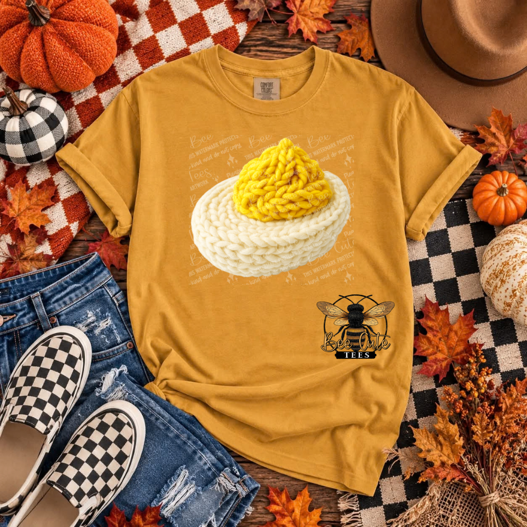 A mustard yellow graphic t-shirt featuring a crocheted egg design and a bee logo, styled in a flat lay with jeans, checkered shoes, pumpkins, and autumn leaves.