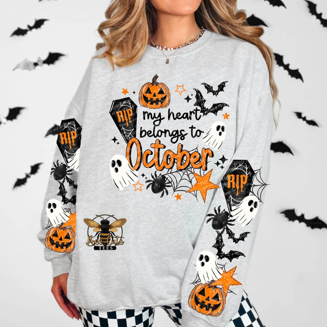 A woman wearing a light gray long-sleeve sweatshirt with a Halloween graphic print that reads 'my heart belongs to October' in orange, featuring pumpkins, bats, ghosts, spiders, and tombstones. The background is white with black bat silhouettes.
