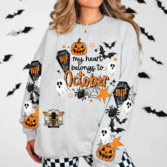 A woman wearing a light gray long-sleeve sweatshirt with a Halloween graphic print that reads 'my heart belongs to October' in orange, featuring pumpkins, bats, ghosts, spiders, and tombstones. The background is white with black bat silhouettes.