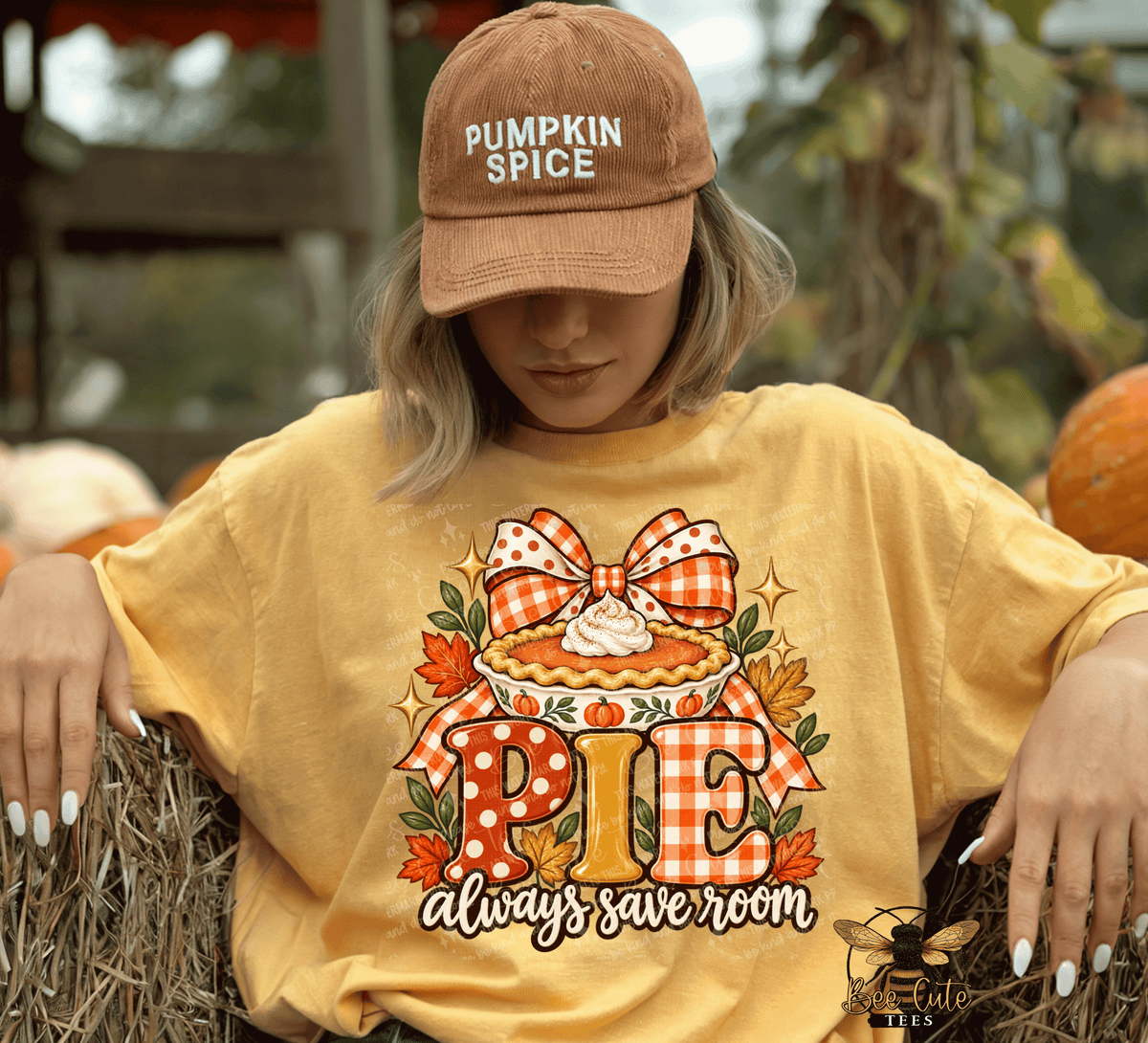 A woman wearing a tan 'Pumpkin Spice' baseball cap and a yellow graphic t-shirt sitting on a hay bale at an outdoor autumn-themed event. The t-shirt features a pie graphic with the text 'PIE always save room'.