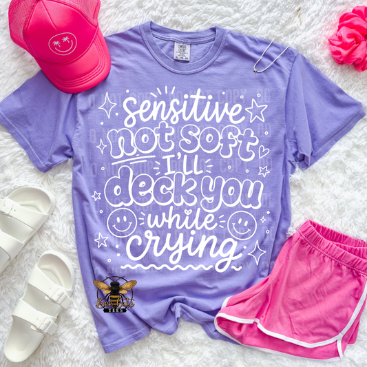 A purple graphic T-shirt laid flat on a white faux-fur surface, styled with a pink cap, white slide sandals, pink shorts, and a small bee logo. The shirt has large white decorative text reading 'Sensitive not soft I'll deck you while crying' with smile faces and star accents.