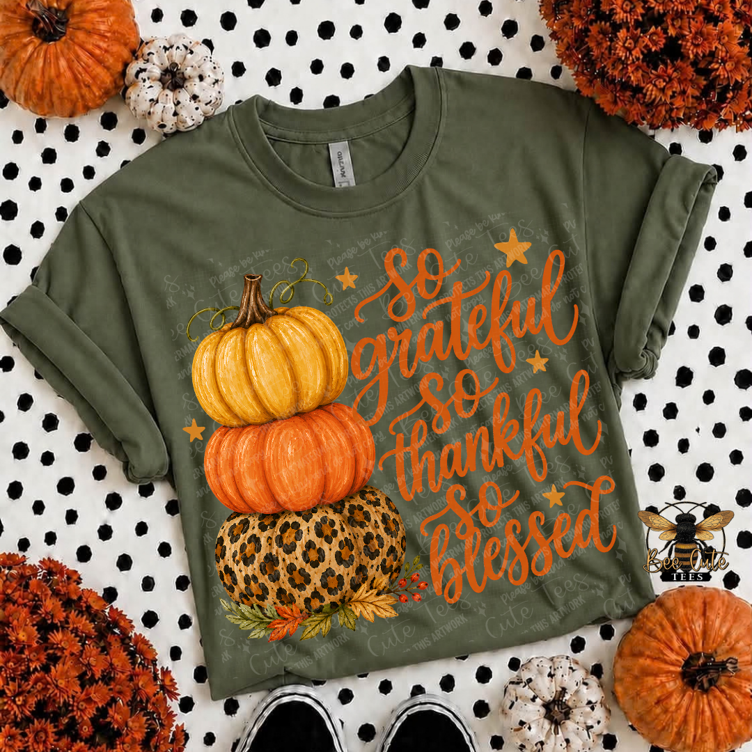 An olive green short-sleeve t-shirt with a 'So grateful so thankful so blessed' graphic featuring stacked pumpkins and a leopard print pumpkin, styled in a flat lay with autumn props.