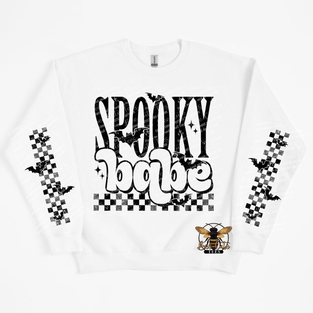 A white crewneck sweatshirt featuring a 'Spooky Babe' graphic on the front with black bats, a checkerboard pattern, and a small bee logo near the lower right front. The sleeves also feature black and white checkerboard patterns with black bats.