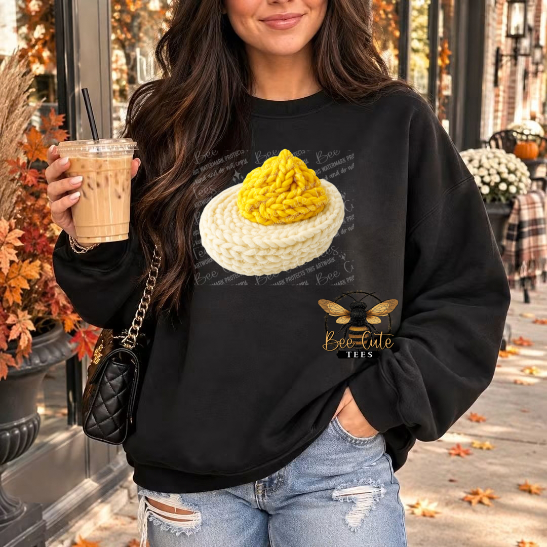 A woman wearing a black crewneck sweatshirt with a graphic of a yellow cheese ball on a white plate and a bee logo that reads 'Be Cute Tees'. She is holding an iced coffee and wearing distressed blue jeans in an outdoor autumn setting.