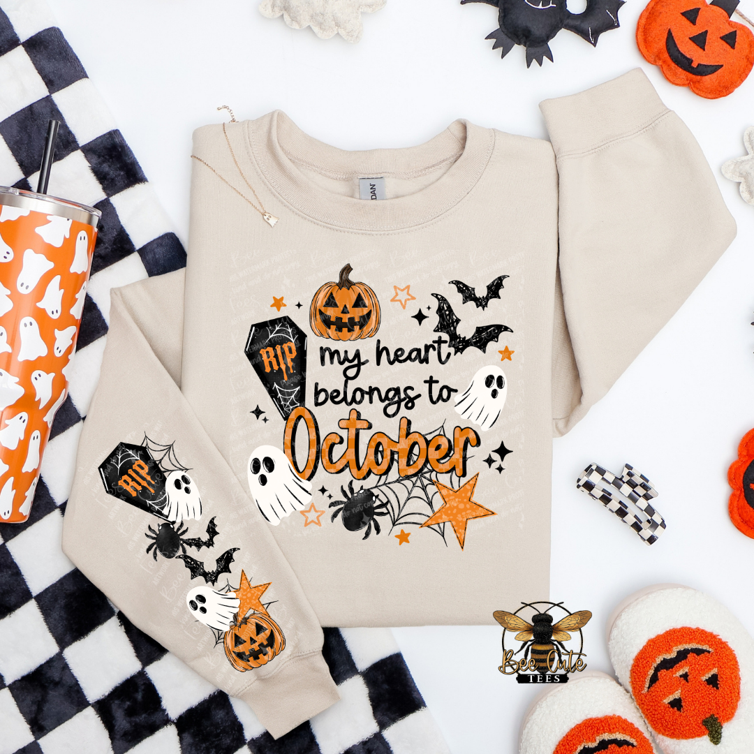 A beige long-sleeve sweatshirt with a Halloween-themed graphic print reading 'my heart belongs to October' in orange and black, surrounded by pumpkins, bats, ghosts, spiders, and stars. The sweatshirt is styled in a flat lay with matching printed accessories and seasonal props on a white background.