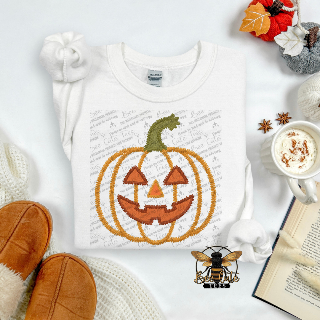 A folded white long-sleeve shirt with a large embroidered orange jack-o'-lantern graphic and a green stem, styled in a cozy flat lay with a mug of hot cocoa, knit socks, a book, and autumn leaves.