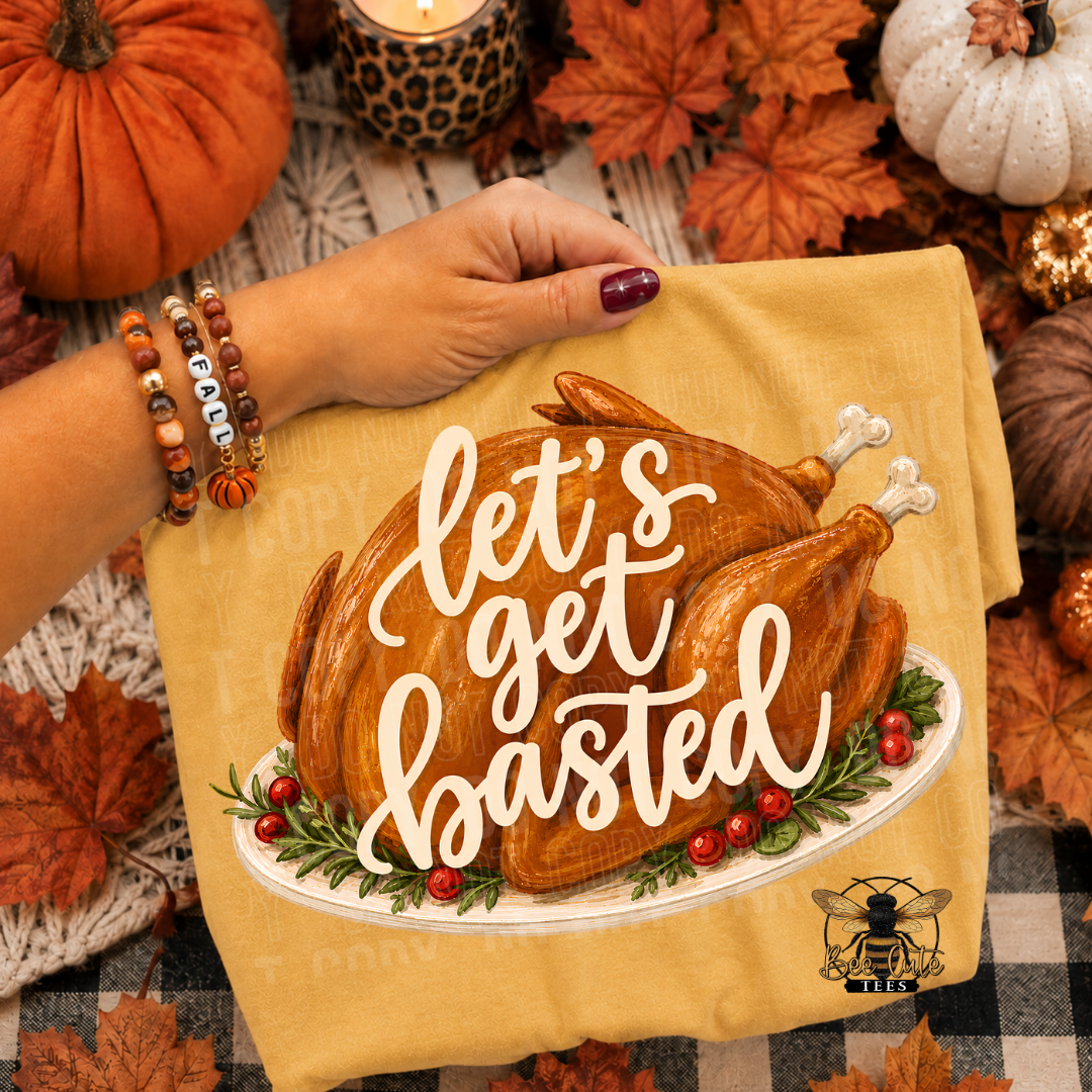A folded mustard yellow t-shirt, featuring a graphic of a roasted turkey on a platter with the phrase 'let's get busted' in white script. The fabric is held by a hand wearing a beaded bracelet, set against a rustic autumn background with pumpkins, fall leaves, and a candle.