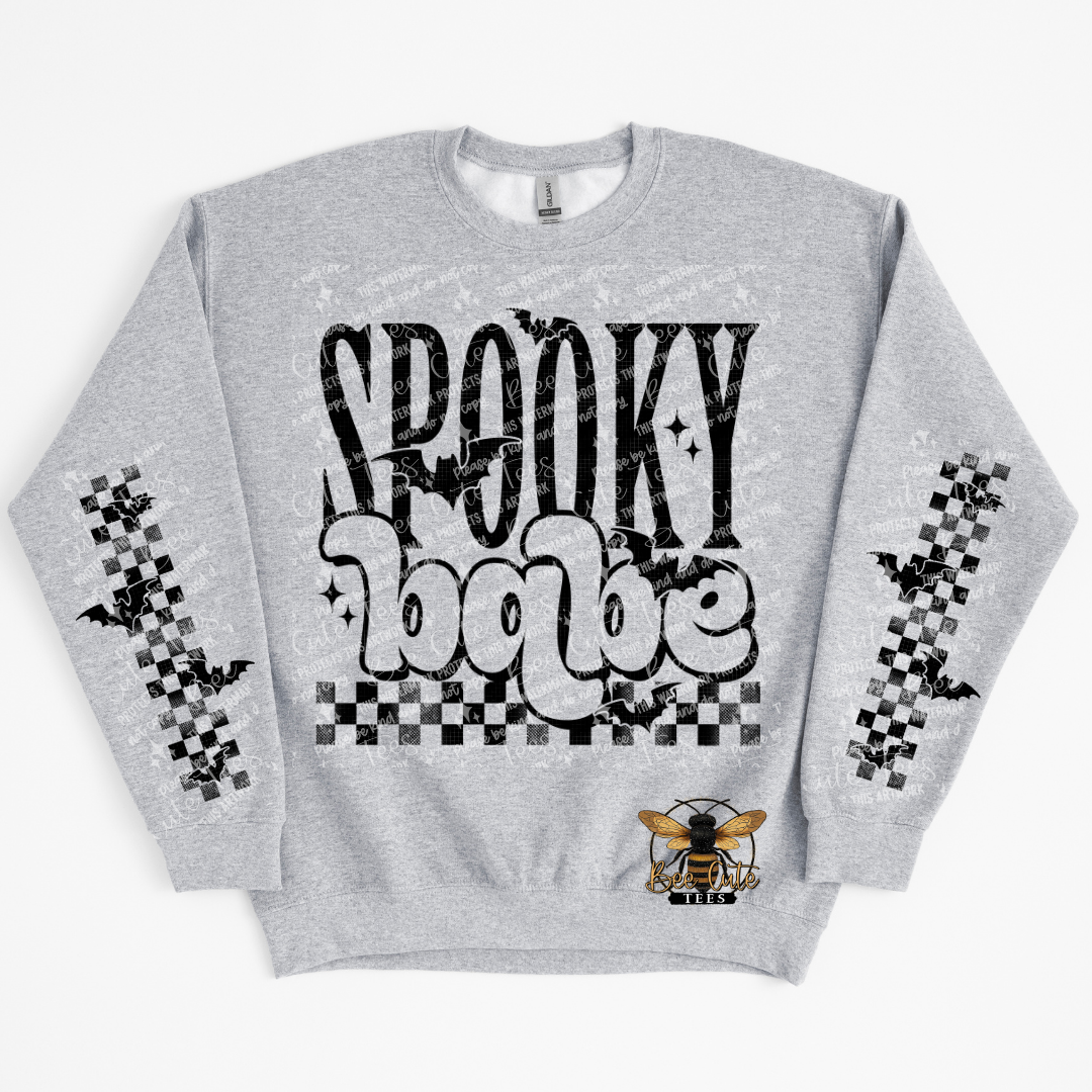 A light heather gray crewneck sweatshirt featuring a 'Spooky Babe' graphic on the front with black and white bubble lettering, checkered bat motifs on the sleeves, and a small bee logo near the lower right front.
