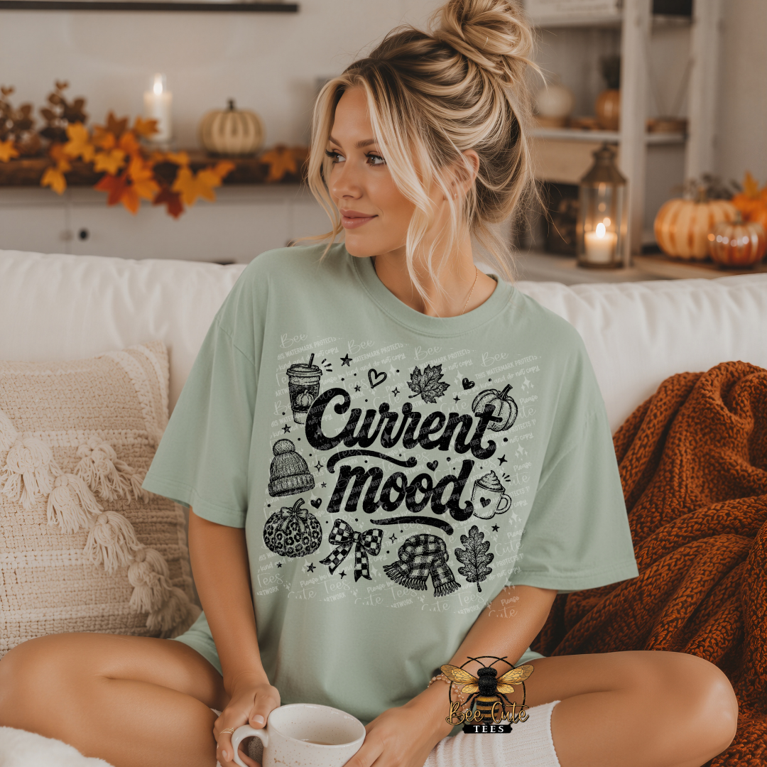 A woman sitting on a white couch wearing a sage green graphic t-shirt with 'Current mood' written in black script surrounded by fall-themed illustrations like pumpkins, leaves, and a beanie. The background shows a cozy living room setting with autumn decorations.