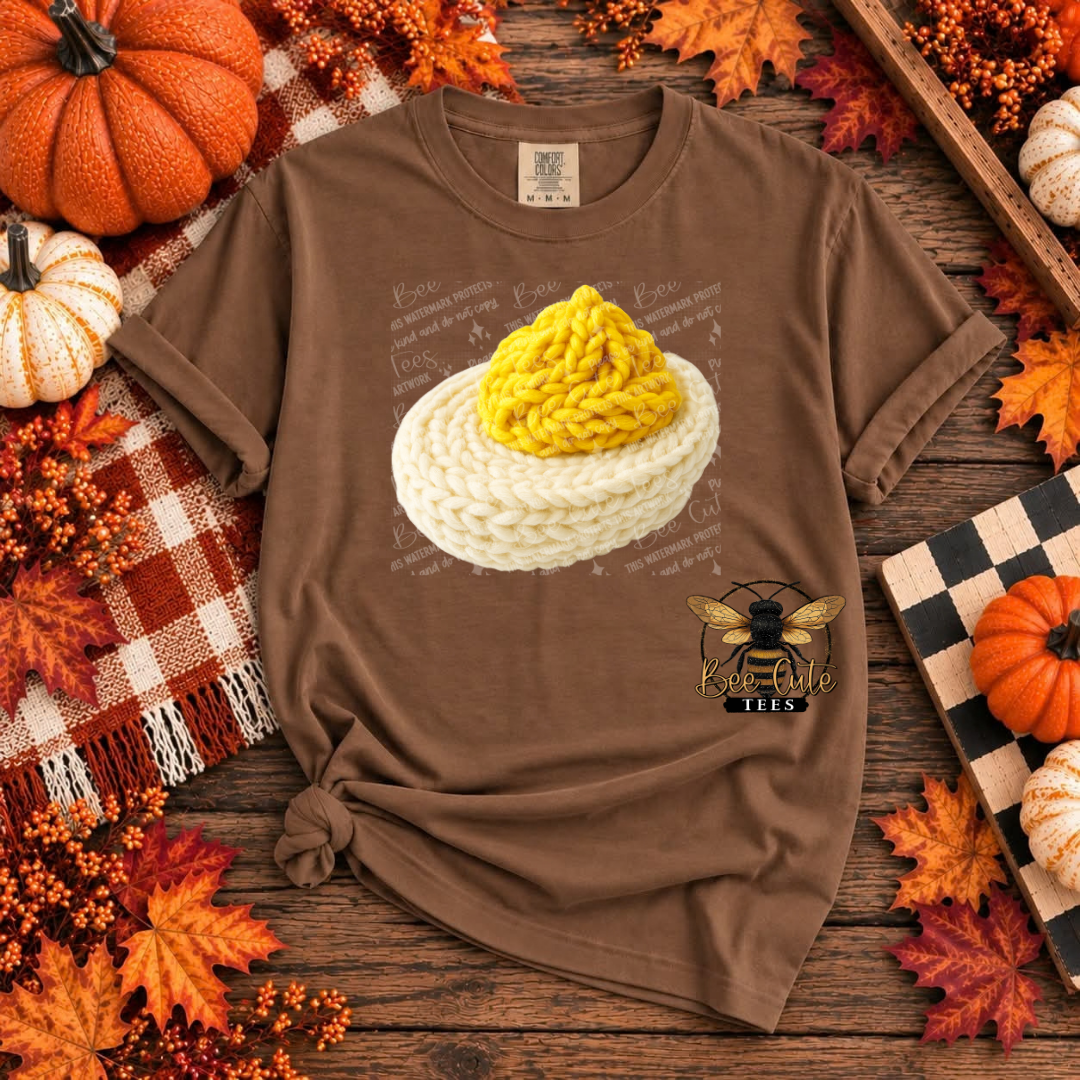 A brown short-sleeve graphic T-shirt laid flat on a rustic wooden surface surrounded by autumn decorations. The shirt features a printed design of a deviled egg, with a bee logo and the text 'BEE CUTE TEES' below.