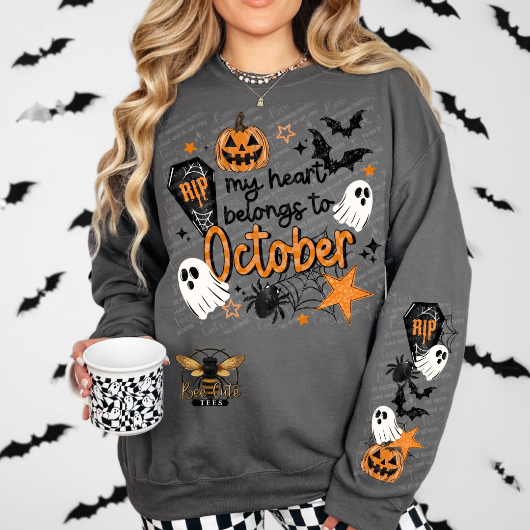 A woman wearing a dark gray crewneck sweatshirt with a Halloween graphic that reads 'my heart belongs to October' in orange, featuring pumpkins, ghosts, spiders, and bats. She is holding a white mug with a black and white pattern, set against a white background with a black bat print.
