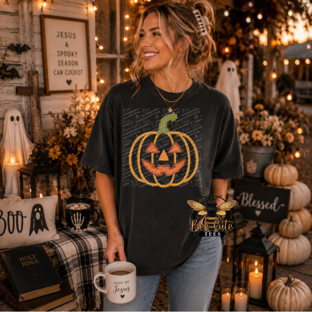 A woman wearing a black graphic t-shirt with a pumpkin design, standing in a cozy, decorated Halloween-themed room. She is holding a white mug and smiling, with pumpkins, candles, and autumn decorations in the background.