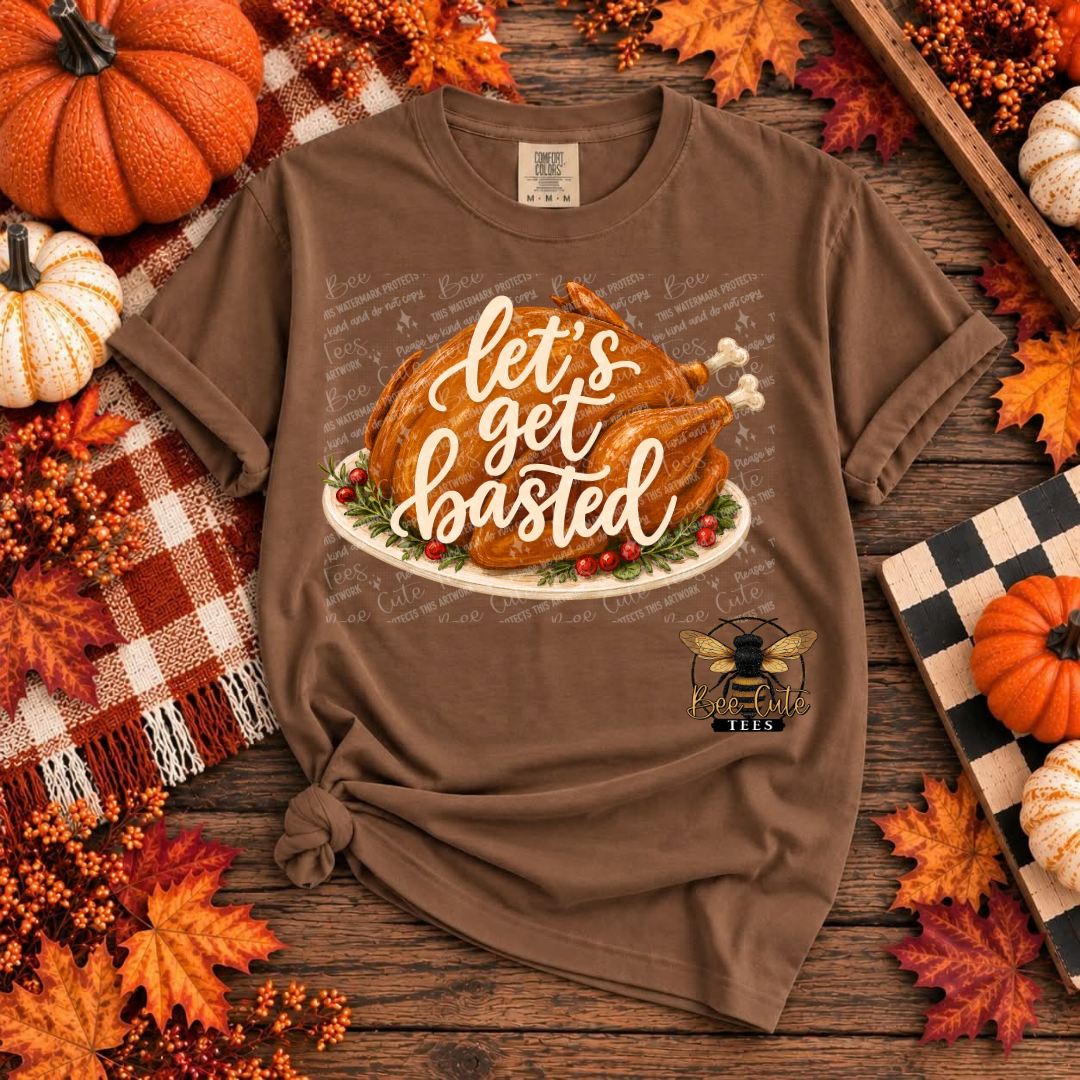 A brown short-sleeve graphic T-shirt laid flat on a rustic wooden surface surrounded by autumn decorations. The shirt features a large Thanksgiving-themed print with a roasted turkey on a platter and the phrase “let’s get basted” in white script.