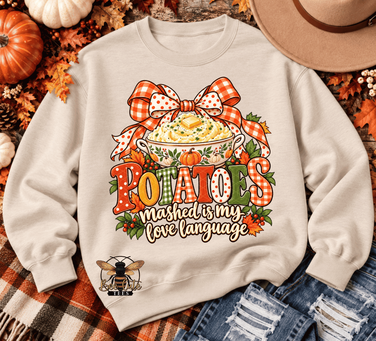 A beige crewneck sweatshirt laid flat with a large autumn-themed potato graphic on the front. The design features a bowl of mashed potatoes, a red-and-white gingham bow, the word 'POTATOES' in colorful letters, and the phrase 'mashed is my love language.