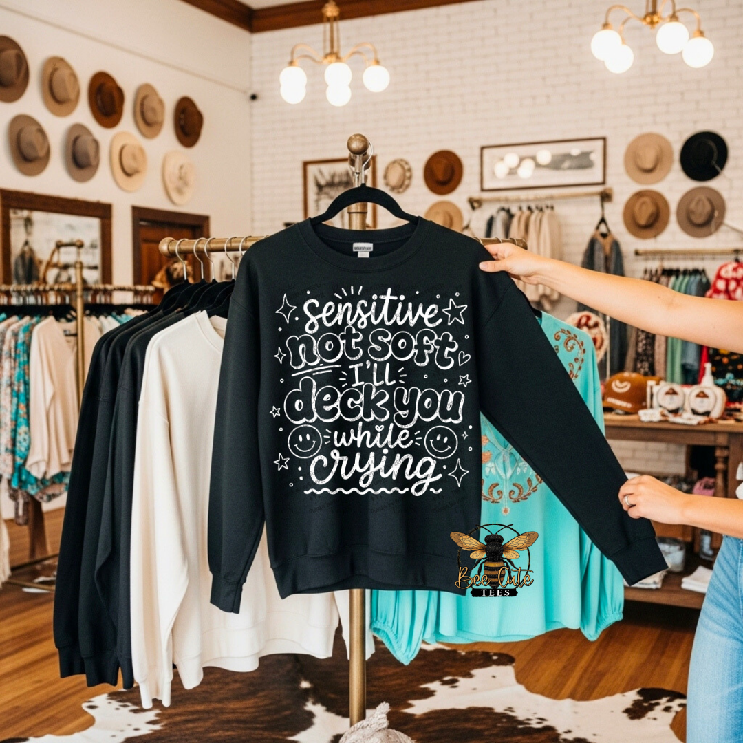 A person holds up a black crewneck sweatshirt on a hanger in a retail clothing store. The sweatshirt features white hand-drawn style text that reads 'sensitive not soft I'll deck you while crying' with doodled stars, smiley faces, and a small bee logo near the bottom right.