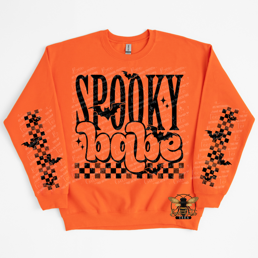 An orange crewneck sweatshirt featuring a 'Spooky Babe' graphic on the front with black bats, checkered patterns on the sleeves, and a bee logo on the lower right front.
